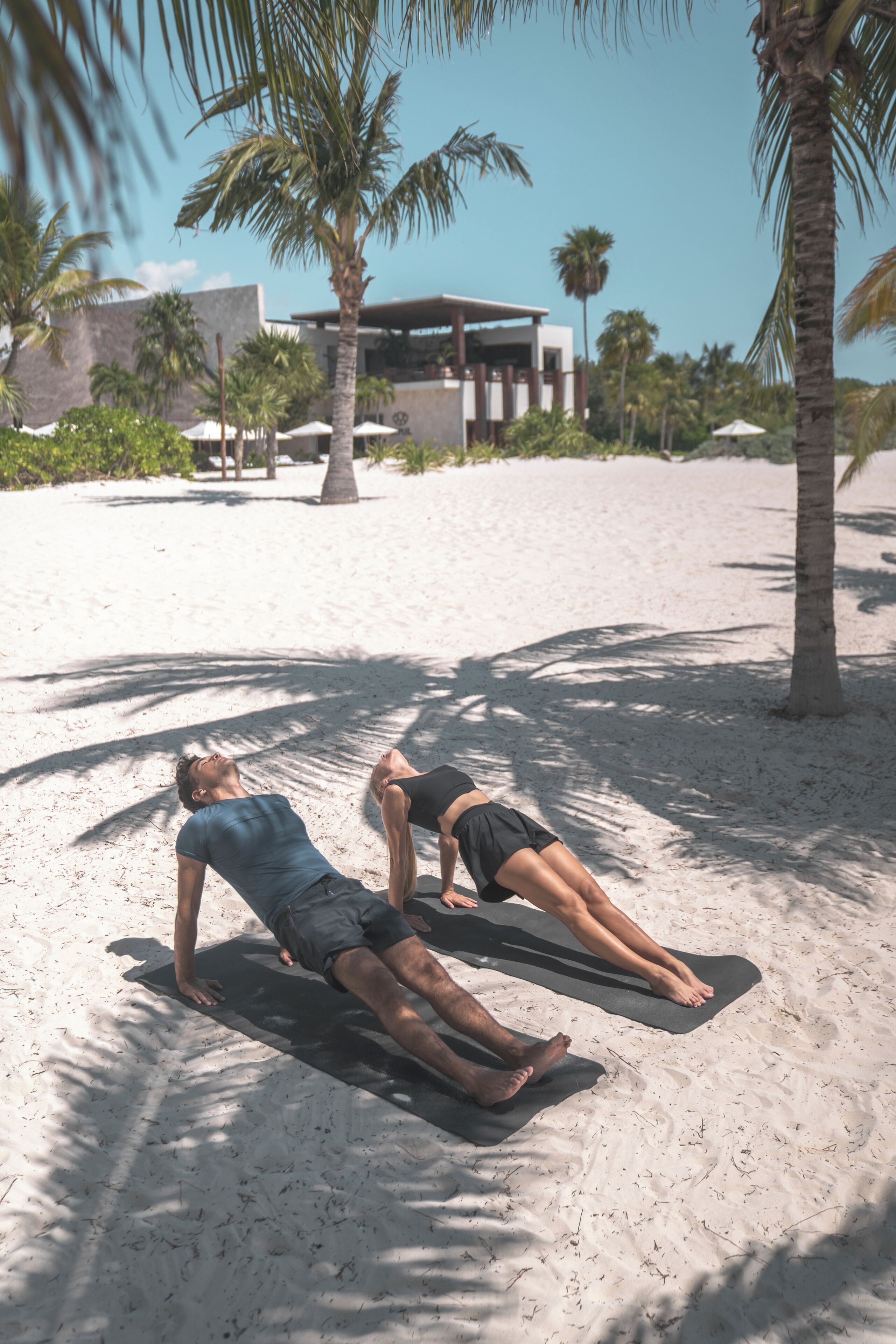 Zwei Personen üben Yoga auf Yogamatten an einem sandigen Strand, im Hintergrund sind Palmen und ein Gebäude zu sehen.