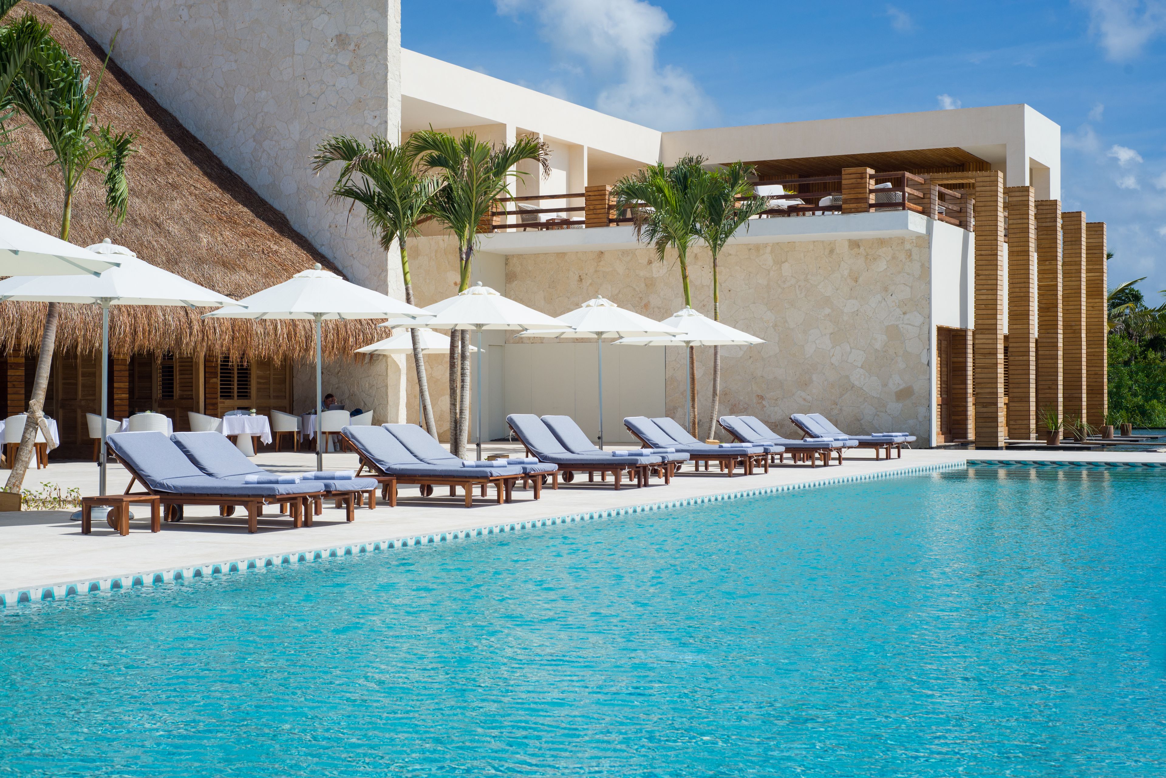 A poolside area with lounge chairs and umbrellas, a thatched roof structure, and a modern building with a stone facade.