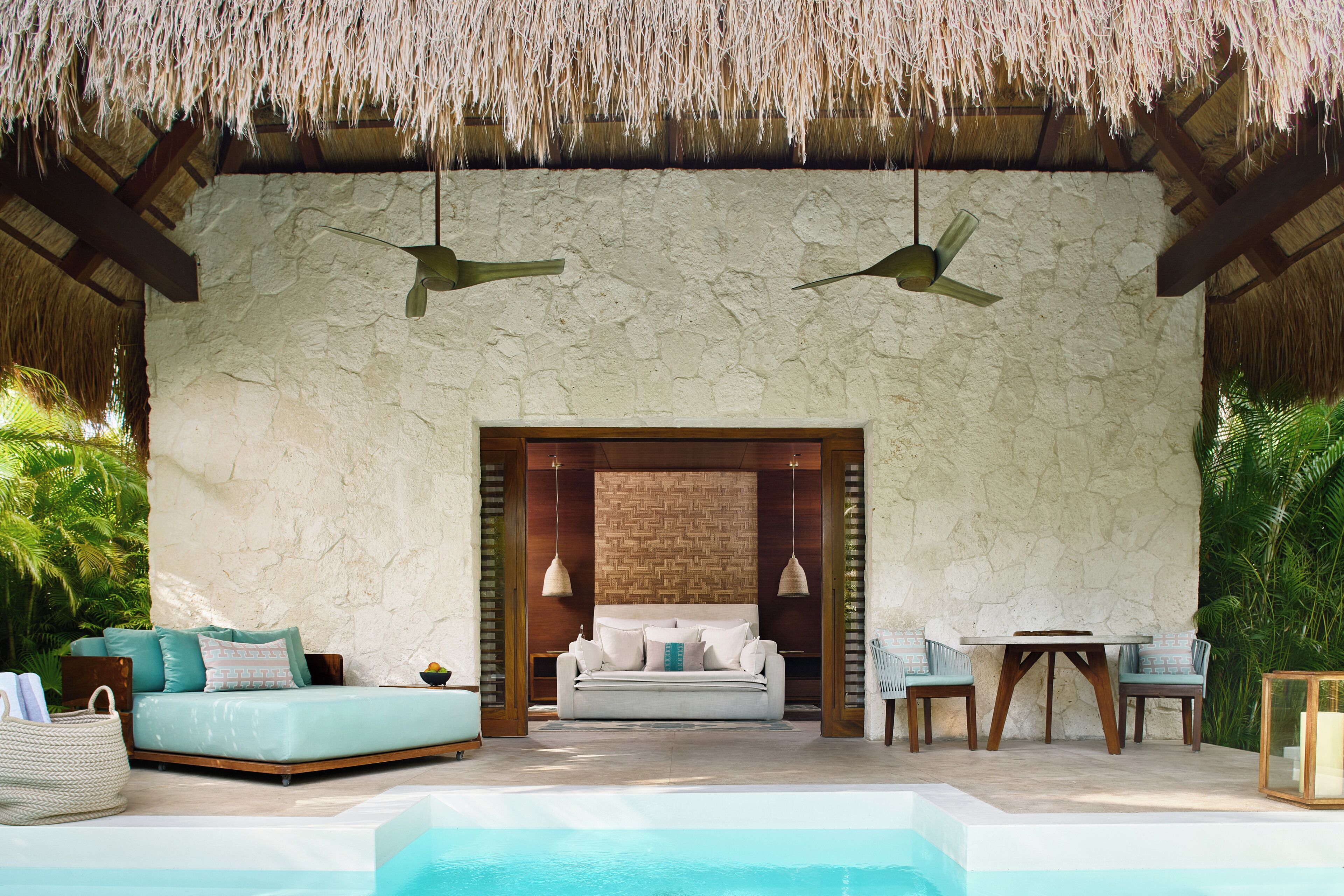 Una habitación con pared de piedra, techo de paja, un sofá, una mesa, sillas y una piscina.
