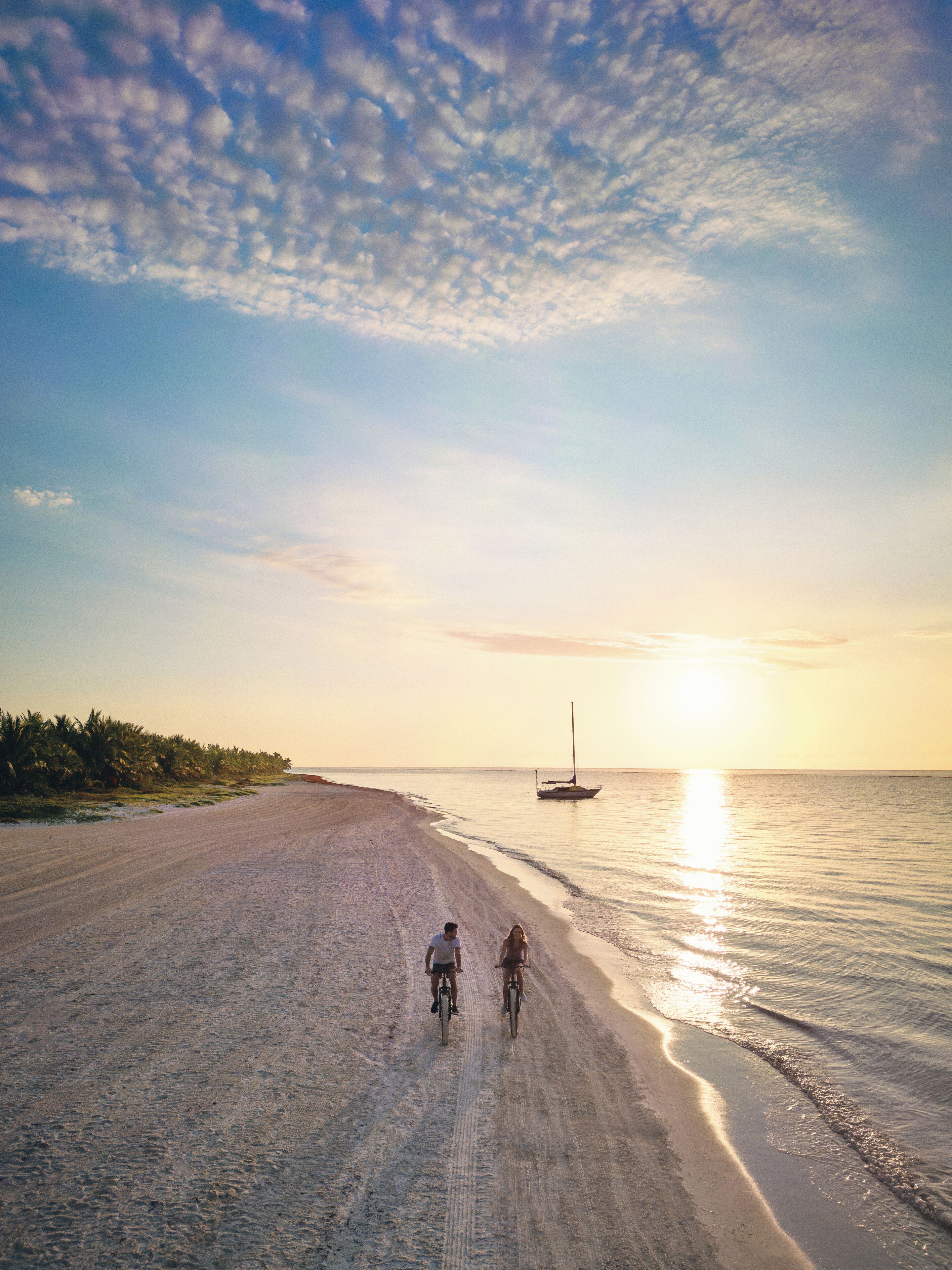 Une plage où deux personnes font du vélo, un voilier à l’horizon et un ciel dégagé.