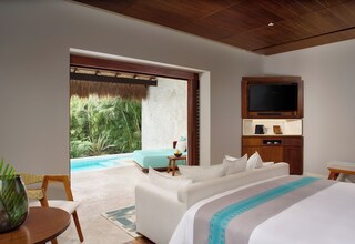 A hotel room with a large bed, a TV mounted on the wall, a seating area with a view of the pool, and a dining table with a plant.