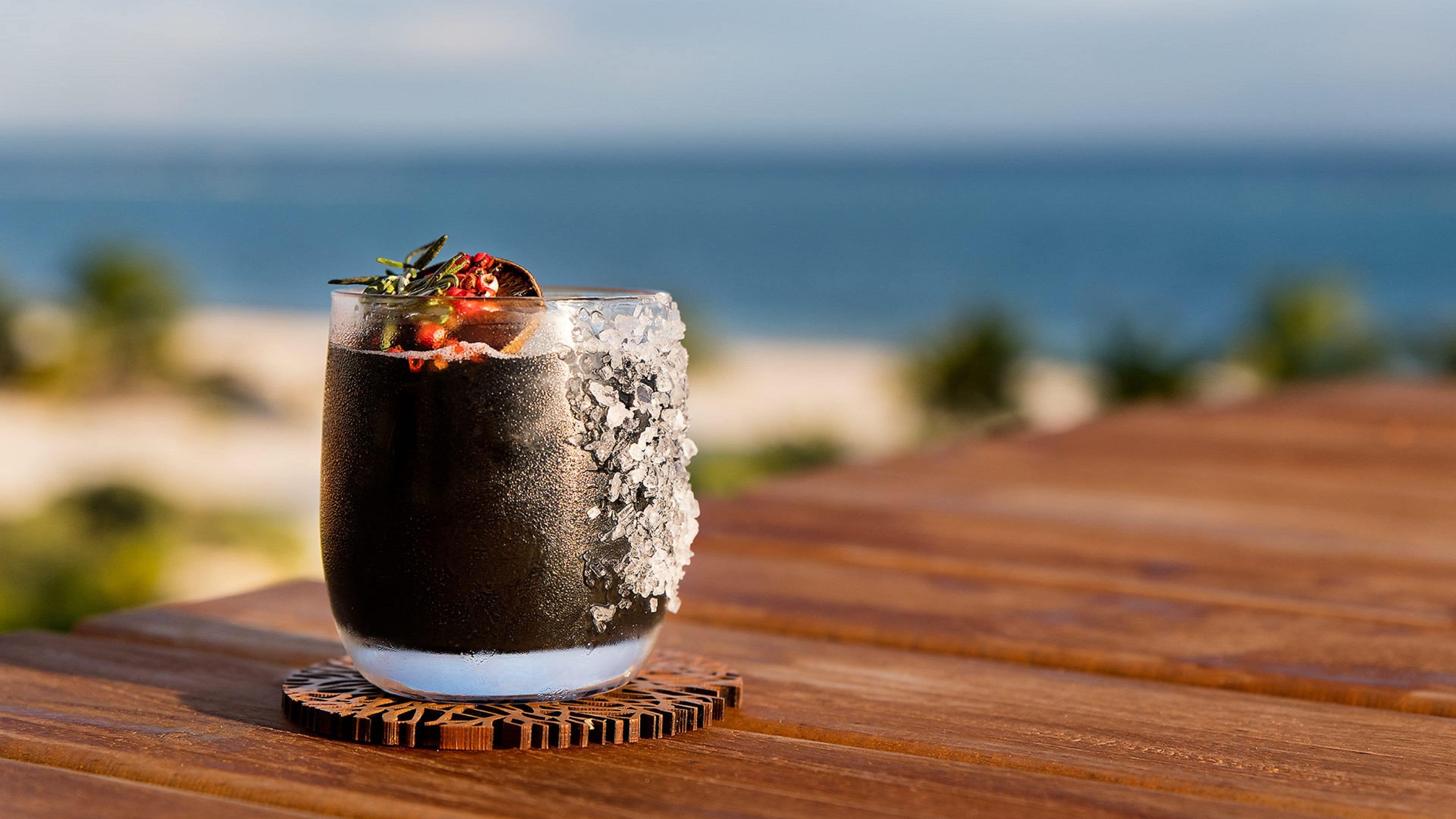 Un vaso de bebida helada con adorno sobre una mesa de madera frente a la playa.