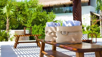 A wooden bench with woven baskets containing towels, set against a backdrop of lush greenery and a thatched-roof building.