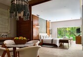 A modern hotel room with a large bed, a dining area with a table and chairs, a fruit bowl, and a view of greenery outside.
