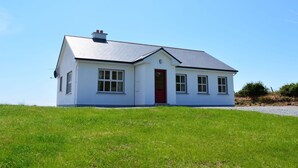 Ein eingeschossiges Haus mit roter Tür, weißen Wänden und grauem Dach, gelegen auf einem grasbewachsen Hügel.