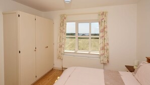 Ein Schlafzimmer mit einem großen Bett, einem Kleiderschrank, einem Fenster mit floralem Vorhang und Blick ins Grüne.