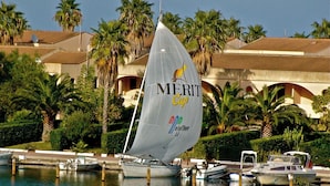 Eine Segelyacht mit 'Merit Cup'-Logo liegt in einer Marina vor Palmen und einem Gebäude im Hintergrund.