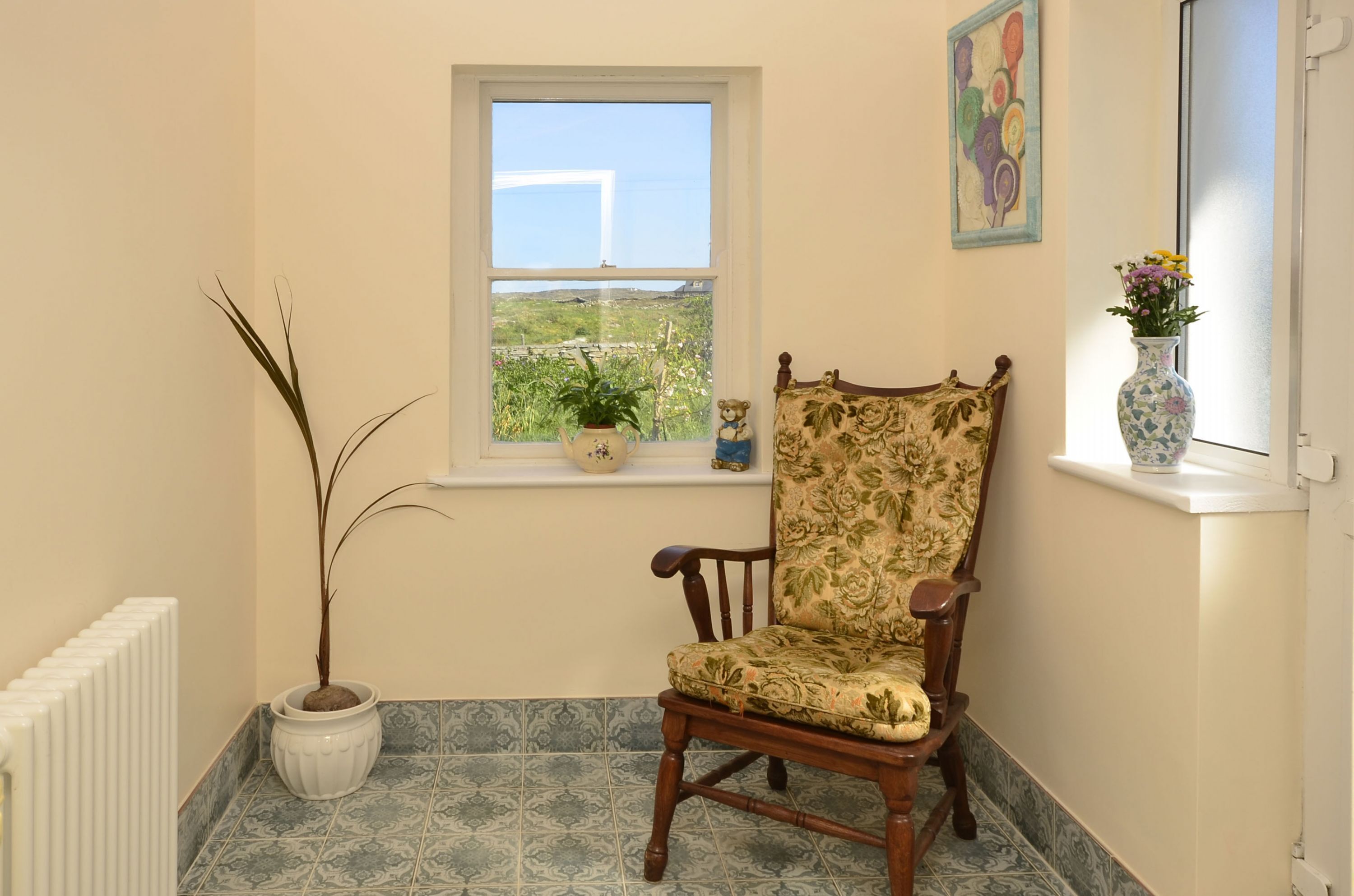 A corner room with a wooden chair, a potted plant, a window with a view, and a vase with flowers.