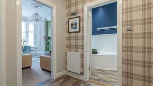 Un couloir avec un radiateur, une porte donnant sur une salle de bain et une vue sur un salon avec un canapé et une lampe.