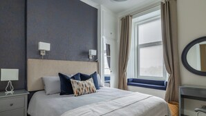 Une chambre à coucher comprenant un lit, des tables de chevet, une commode, une fenêtre avec des rideaux, un miroir et une vue sur l’extérieur.