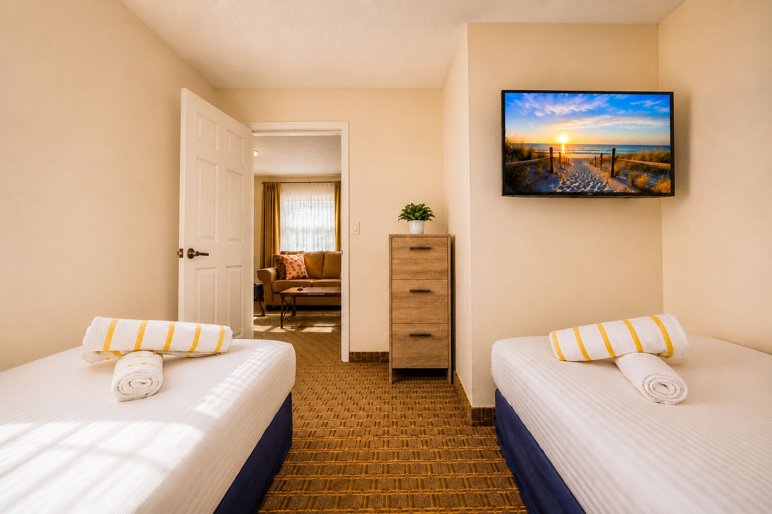 Habitación de hotel con dos camas, un aparador de madera, un cuadro con una escena de playa y vista al área de estar contigua.