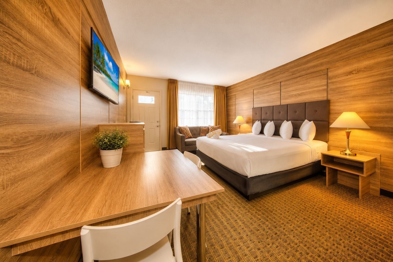 Habitación de hotel con cama, mesita de noche, lámpara, escritorio, silla y una planta en maceta.