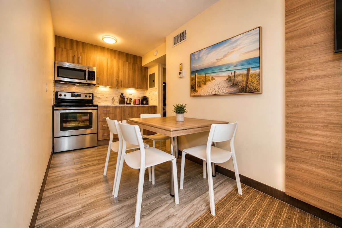 Een compacte keuken met moderne apparaten, een eethoek met witte stoelen en een schilderij met een strandthema aan de muur.