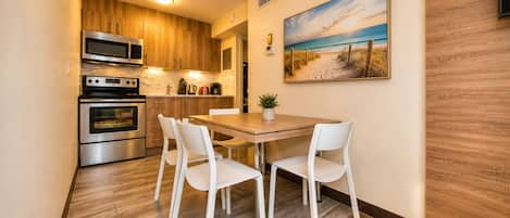 Et kompakt køkken med moderne hårde hvidevarer, et spiseområde med hvide stole og et maleri med et strandmotiv på væggen.