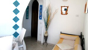 Una habitación con cama, una silla, una mesa, un florero con plantas y un acceso a otra habitación.