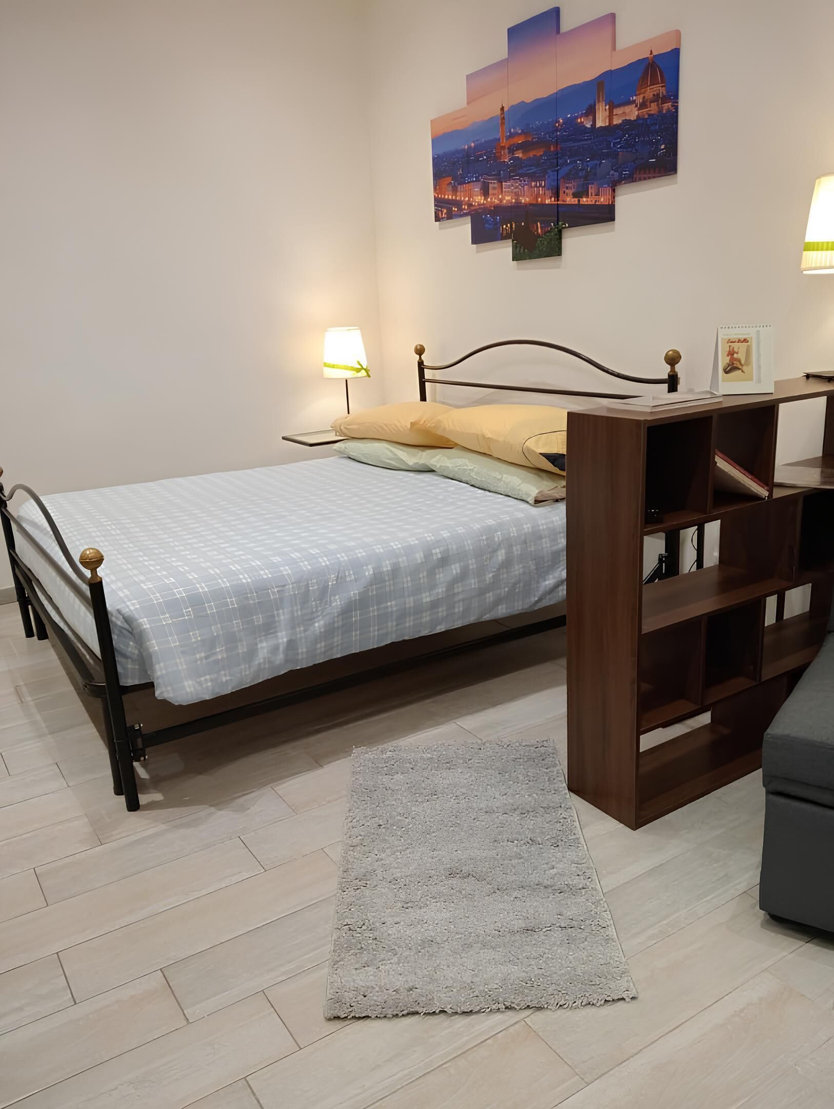A bedroom with a bed, bedside lamps, a nightstand, a bookshelf, a rug, and a cityscape painting on the wall.