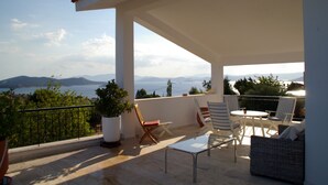 A balcony with a view of the sea and mountains, furnished with chairs and a table.