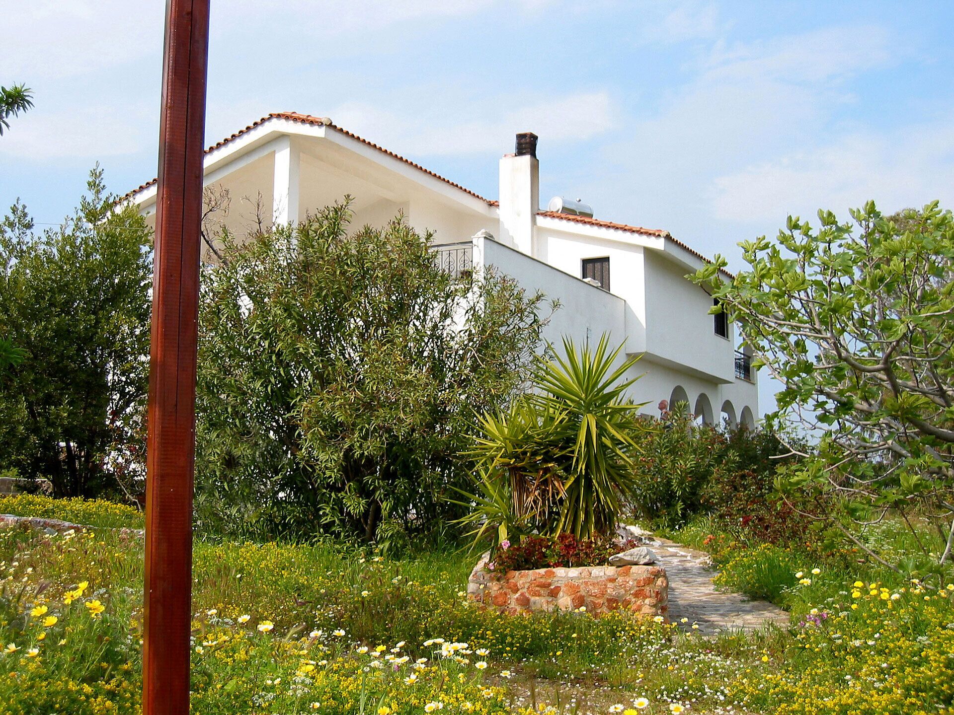 Eine weiße Villa mit einem Ziegeldach, umgeben von üppiger Vegetation und verschiedenen blühenden Pflanzen.