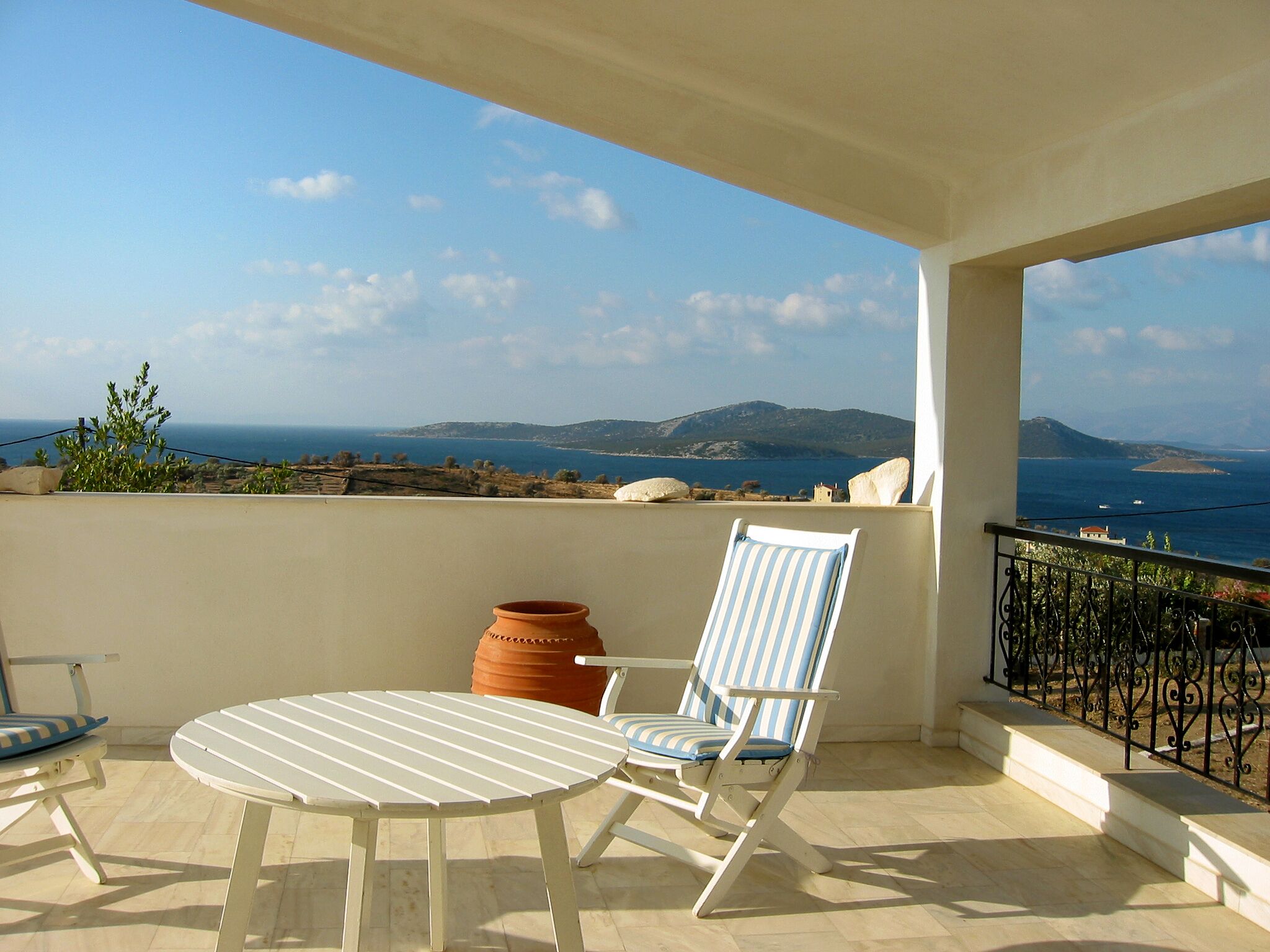 Un balcon avec une table blanche et une chaise à rayures, donnant sur un paysage côtier avec des montagnes et la mer.