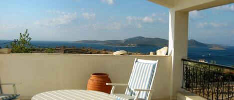Un balcon avec une table blanche et une chaise à rayures, donnant sur un paysage côtier avec des montagnes et la mer.