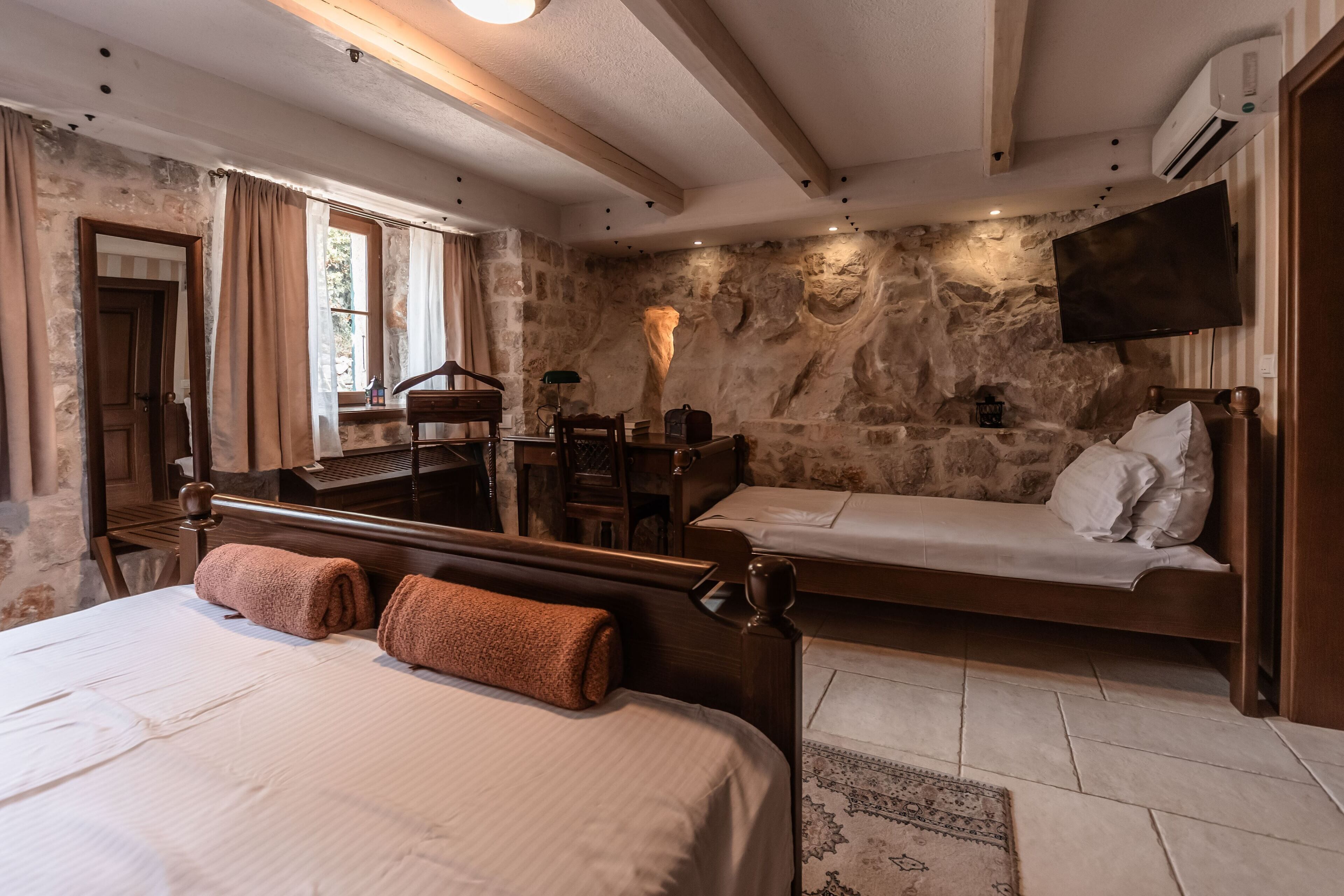 A bedroom with a large bed, a smaller bed, a desk, a chair, and a stone wall.