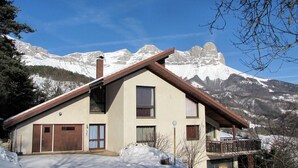 Un chalet à deux étages, doté d’un toit en pente, d’un balcon en bois et offrant une vue sur des montagnes enneigées.