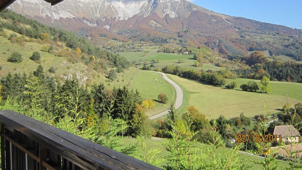 Un paysage de montagne avec une route serpentant à travers une vallée, une terrasse en bois au premier plan et un cachet indiquant la date dans le coin inférieur droit.