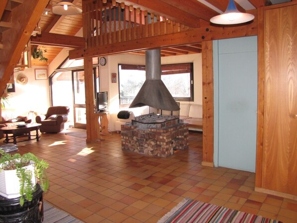 A cozy living room with a stone fireplace, wooden ceiling beams, and a large window. - WILDERNESS PARK VERCORS BEAUTIFUL HOUSE WITH PANORAMIC VIEWS (Saint-Andéol)