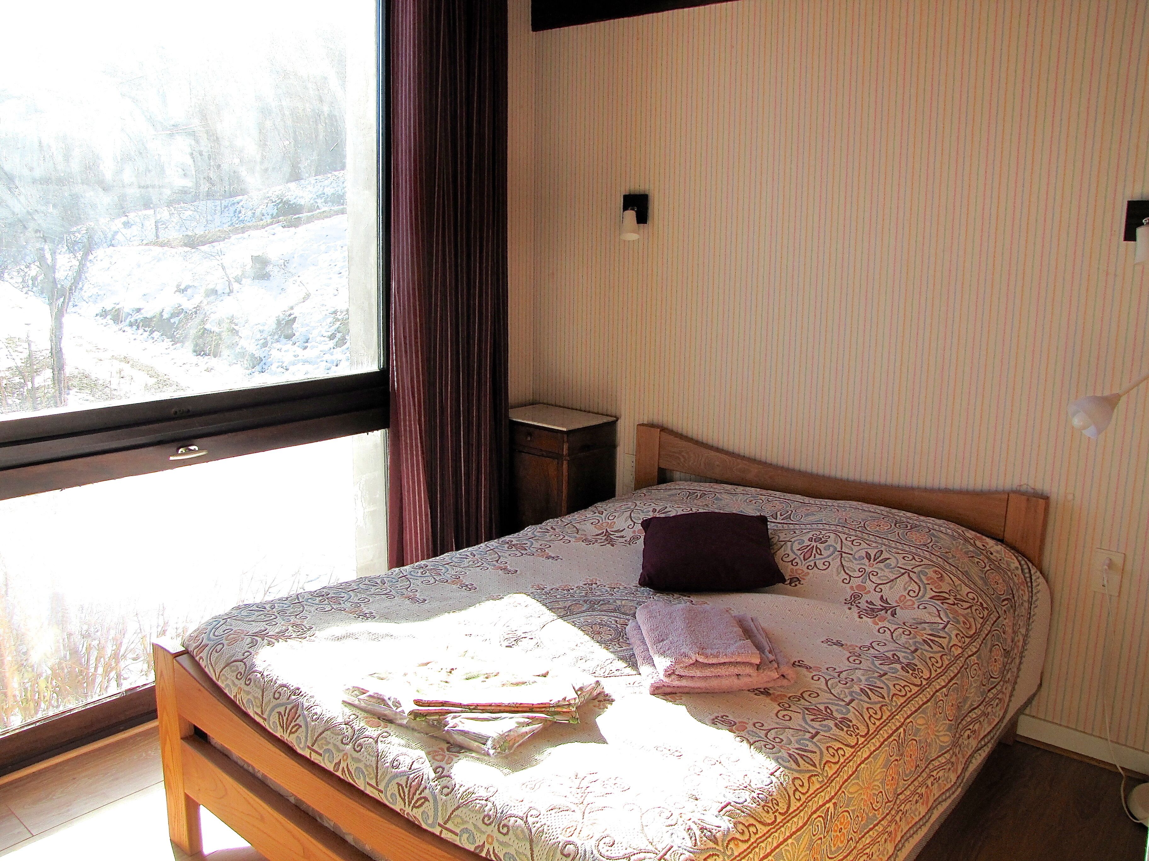 Ein Schlafzimmer mit großem Fenster, einem Bett mit gemustertem Überwurf, einem Nachttisch und einer Wandlampe.