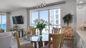 A modern living room with a glass table, wooden chairs, a flat-screen TV, and a large window offering a view of a building and the sea. - Sterling Sands - Gulf front - Sleeps 8 - Three Bedroom - 3 Bath (Destin)