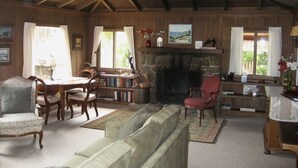 A cozy living room with a stone fireplace, wooden ceiling beams, a sofa, a dining table with chairs, and a bookshelf. - Sweet, Historic Cottage on Carmel Point (Carmel)