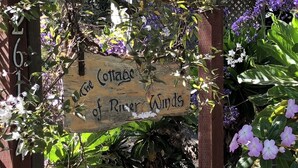 A wooden sign with the text "The Cottage of River Winds" amidst lush greenery and flowers. - Sweet, Historic Cottage on Carmel Point (Carmel)