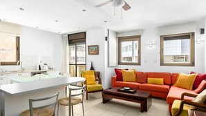 A modern living room with a sofa, chairs, a coffee table, and a kitchen area with a dining table. - MAMAD Cosy Hideaway steps from Beach by FeelHome (Tel Aviv - Jaffa)