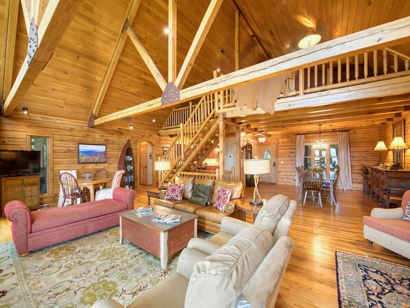 A spacious log cabin interior with a high ceiling, wooden beams, a staircase, a fireplace, and a cozy seating area with a television and books.