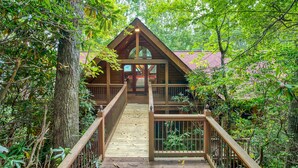A wooden cabin with a wooden deck leading to a glass door entrance, surrounded by dense foliage. - 4 BR Mountain cabin with beautiful long range views in quiet gated community (Maggie Valley)
