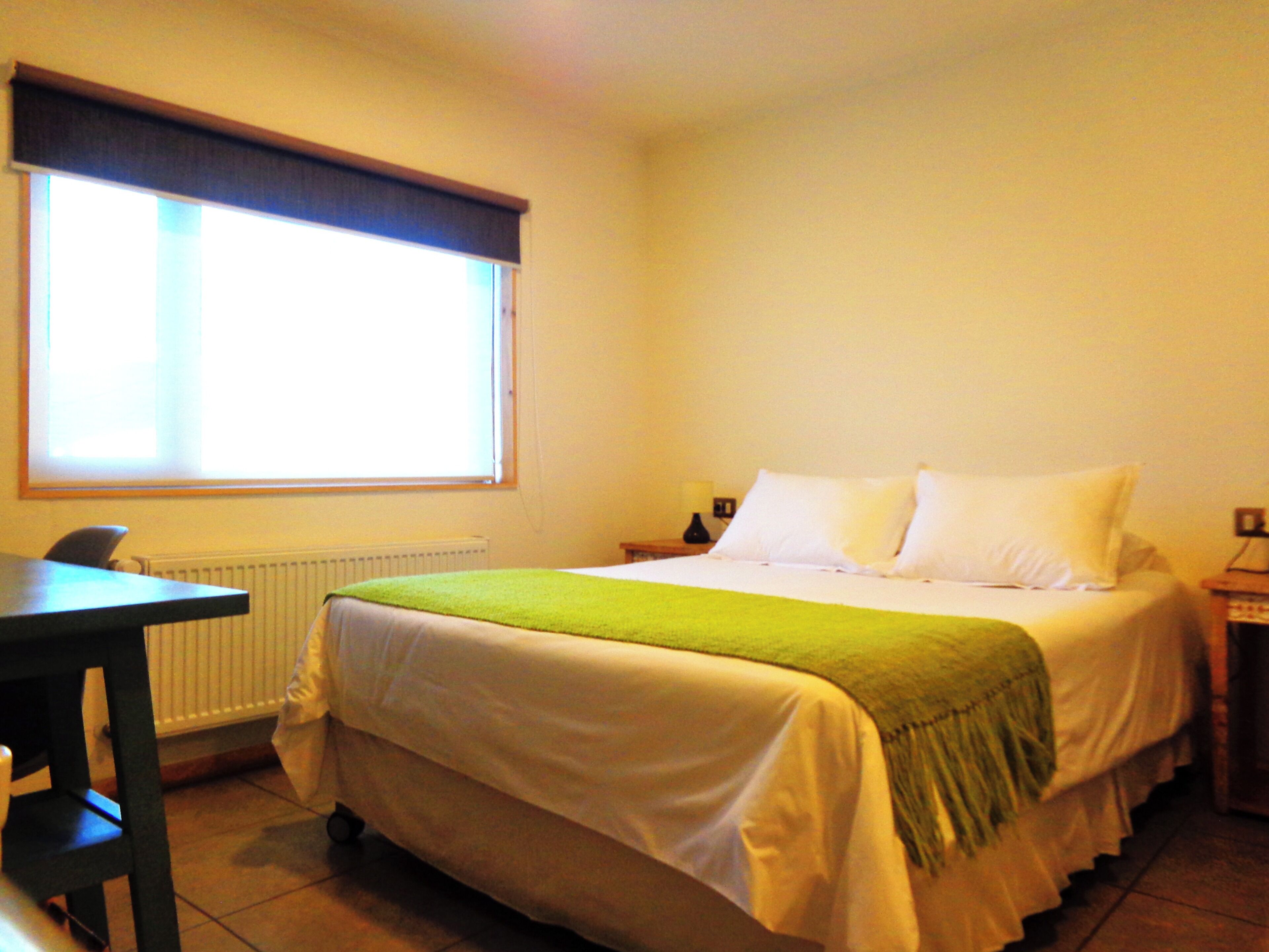 A hotel room with a bed, a chair, a table, a window with blinds, and a radiator.