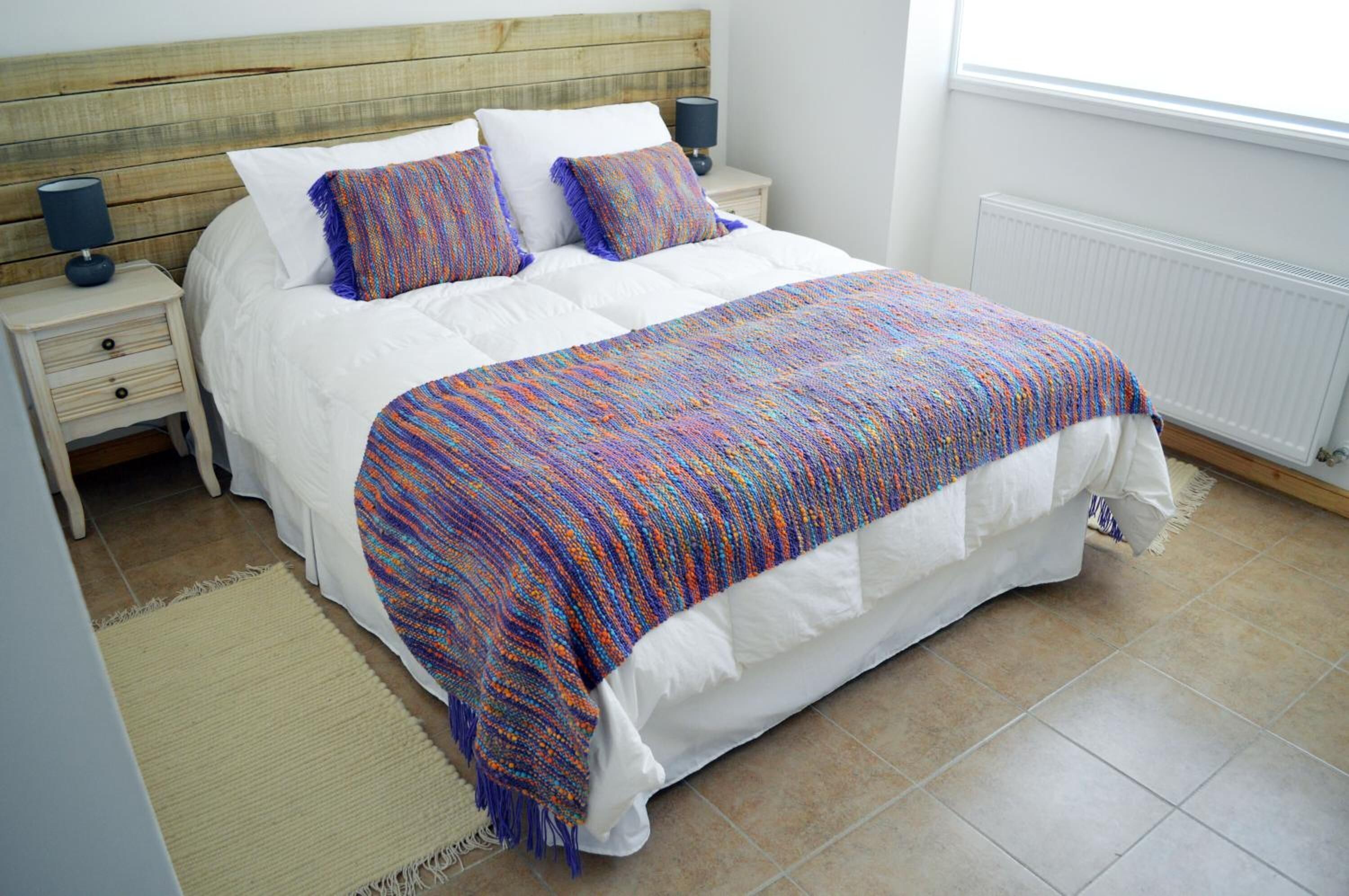 A neatly made bed with a colorful blanket and pillows, a wooden headboard, a bedside lamp, and a radiator.