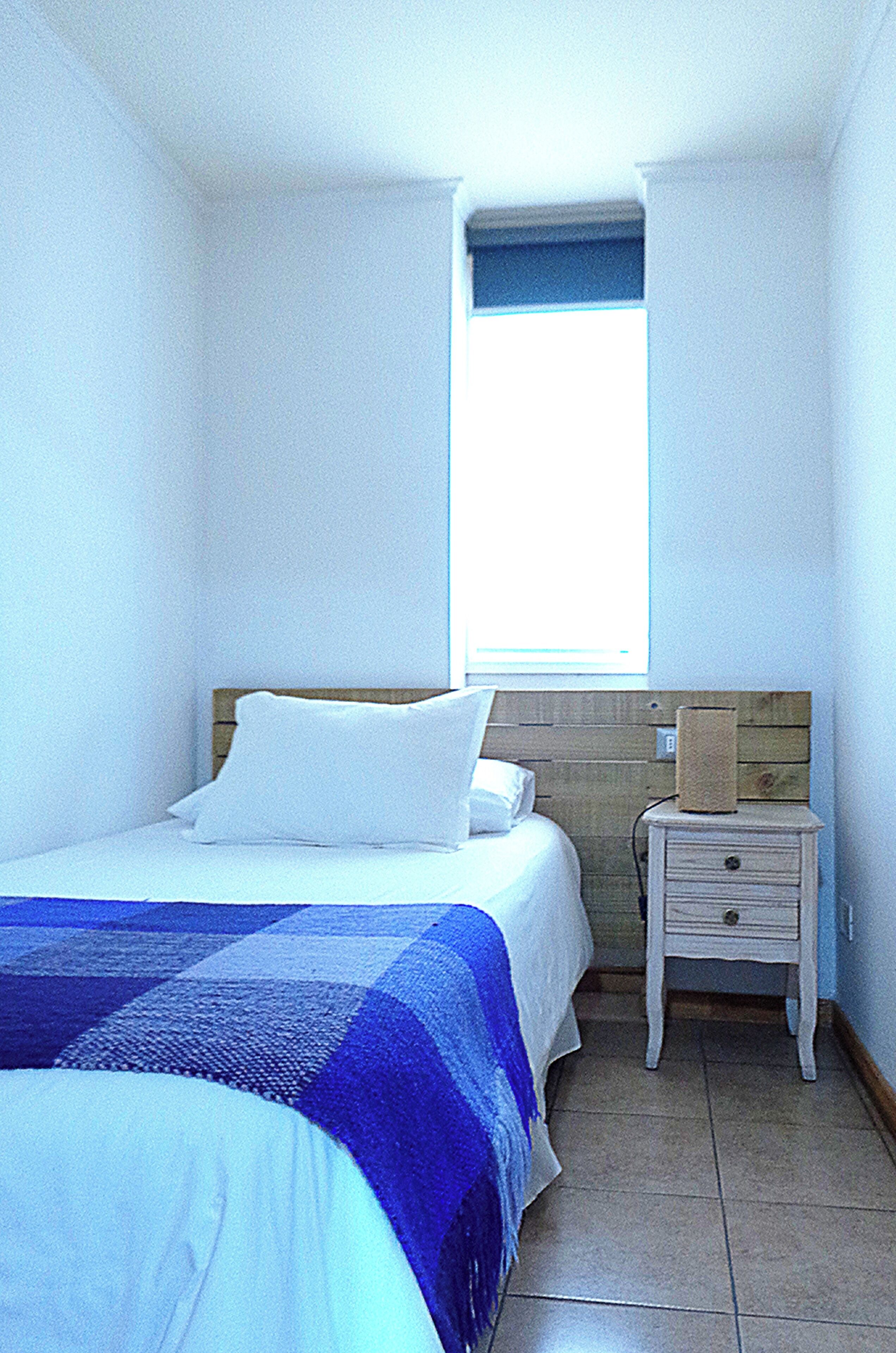 Una habitación pequeña y estrecha con una cama, una mesita de noche y una ventana.