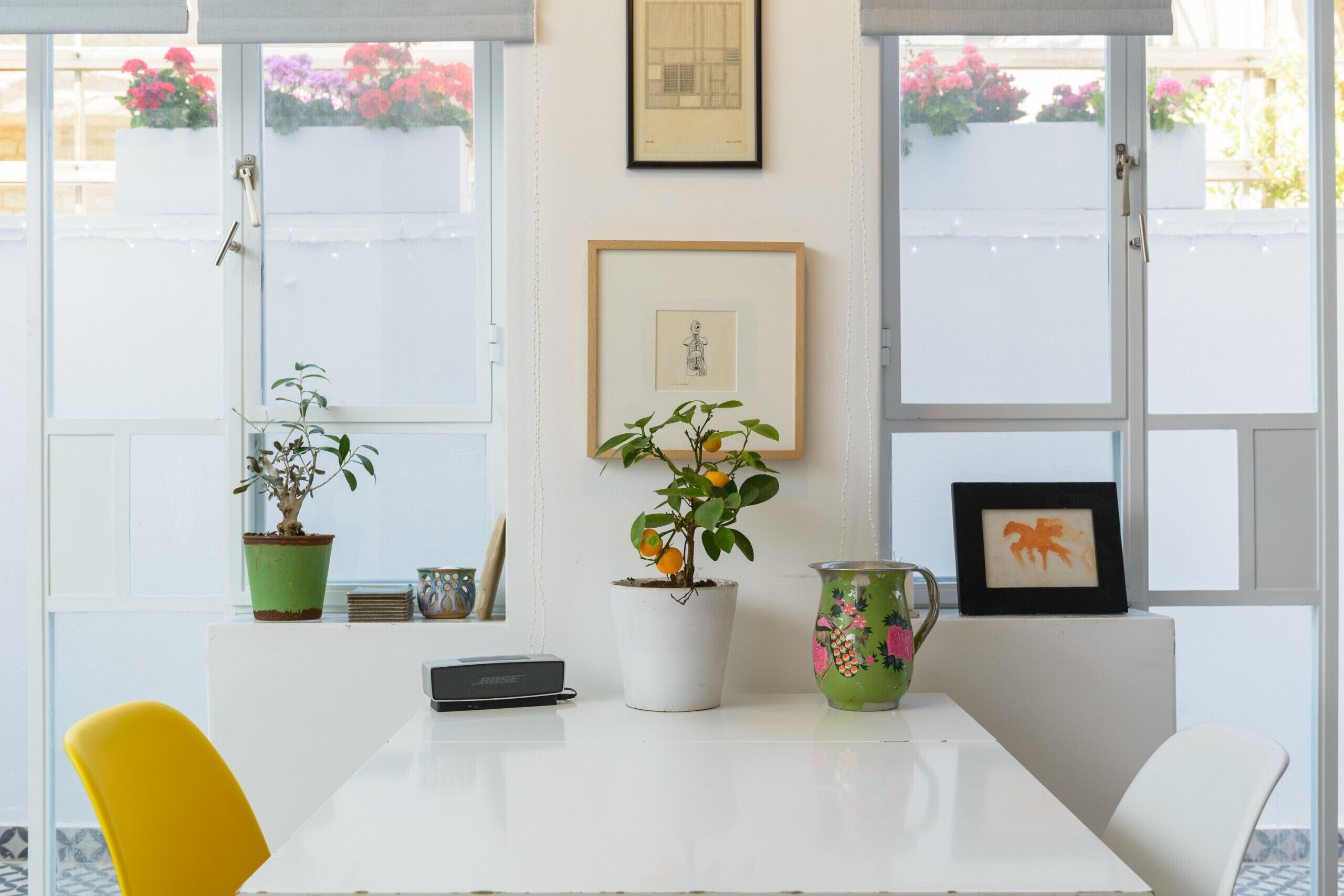A dining area with a white table, chairs, a potted plant, a framed picture, and a vase.