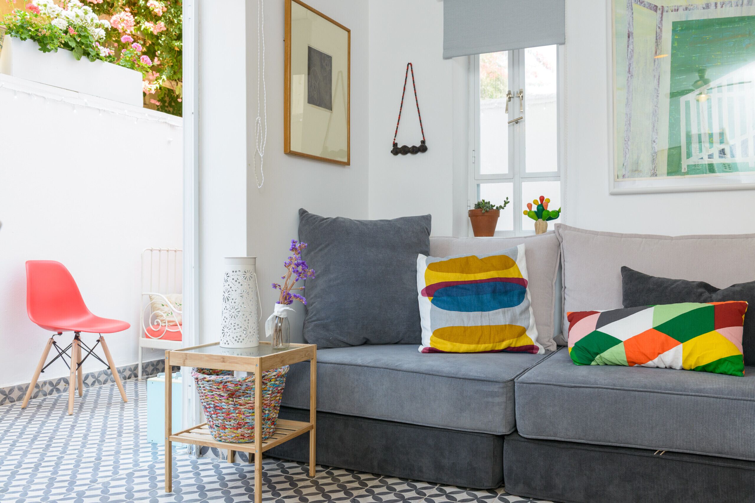 A modern living room with a grey sofa, colorful pillows, a small table with a vase, and a red chair.