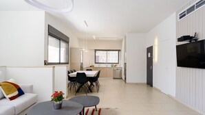 Een moderne woonkamer met een bank, salontafel en een eethoek met stoelen en een tafel. Achtergrond toont een keuken met een raam en een aan de muur bevestigde televisie.