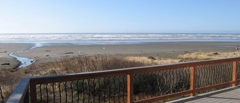 A wooden deck with a railing overlooking a beach with a calm sea and a clear sky.