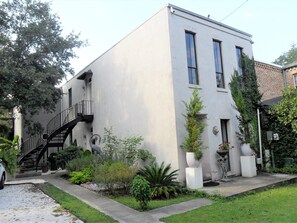 Un bâtiment moderne à deux étages, doté d’une façade blanche, d’un escalier noir et d’un petit jardin agrémenté de diverses plantes et d’un grand bac à fleurs.