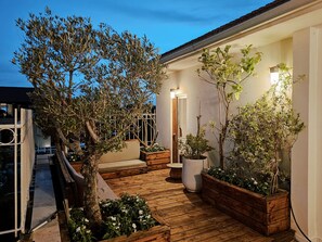 Una terraza de madera con plantas en macetas y un banco, que conduce a un edificio blanco con una puerta y una lámpara.