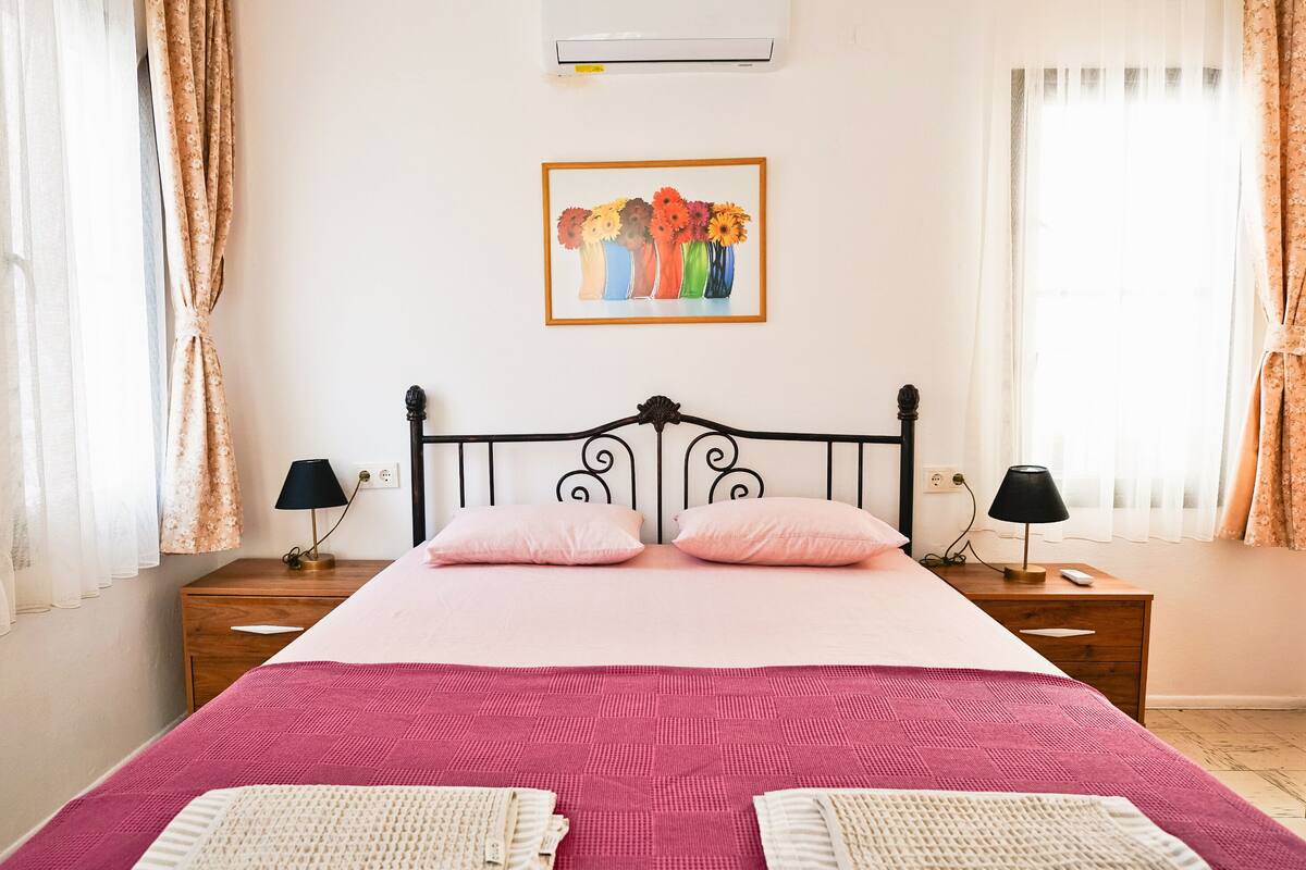 Un dormitorio con cama, dos mesitas de noche, aire acondicionado instalado en la pared, un cuadro con flores y cortinas.