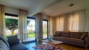 Uma sala de estar com sofá, mesa de centro e um canape. Janelas amplas com cortinas oferecem vista para o verde lá fora.