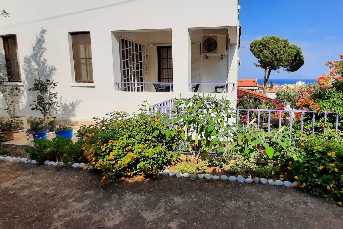 Un edificio blanco con múltiples ventanas y un balcón. Hay plantas en macetas y un pequeño jardín con flores naranjas.