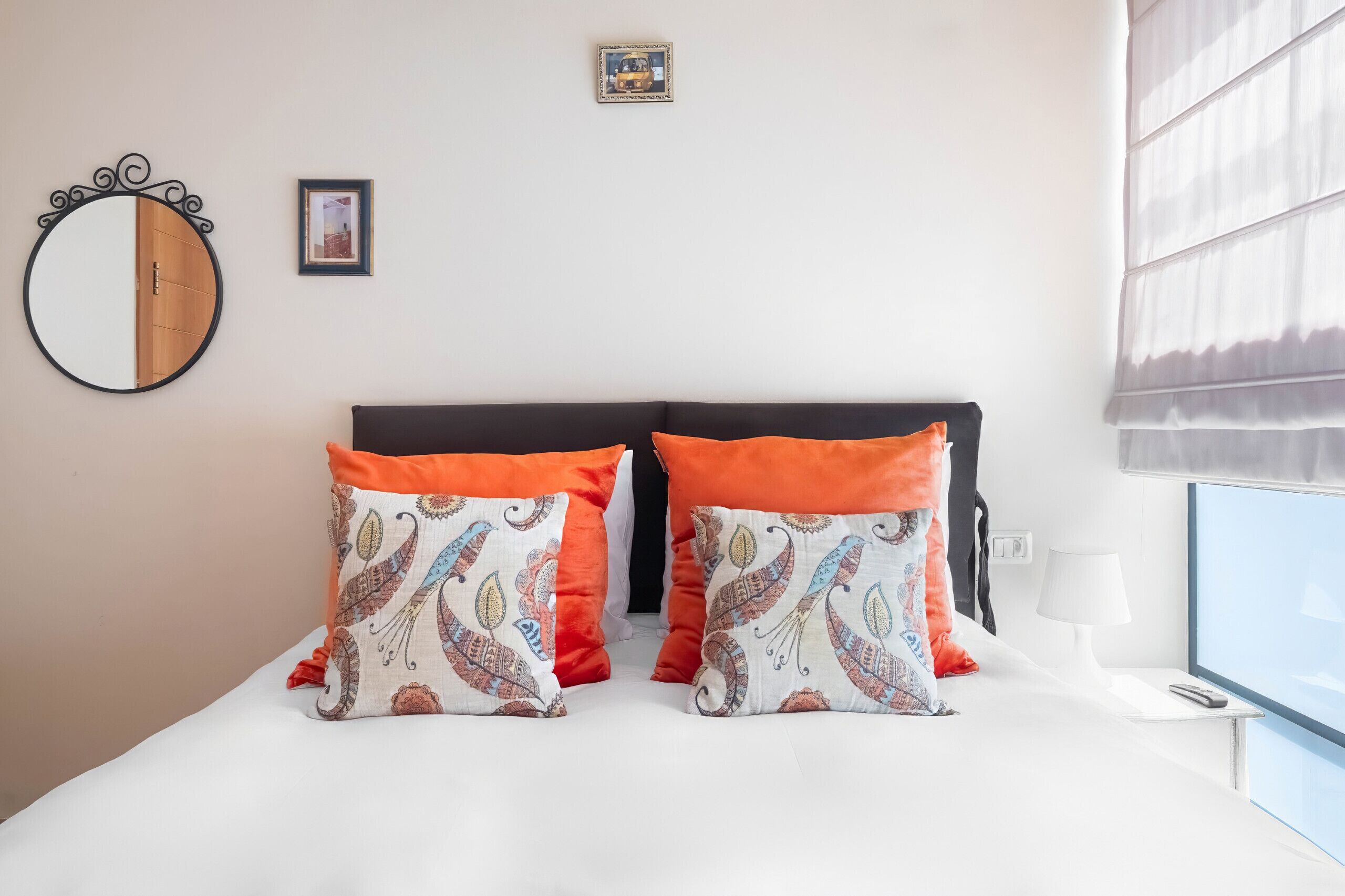 A bedroom with a bed, two decorative pillows, a round mirror, a framed picture, a bedside lamp, and a window with blinds.