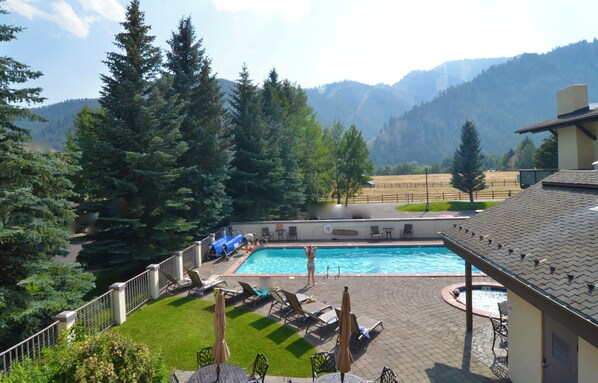 Une piscine entourée de chaises longues et de parasols, elle-même délimitée par une clôture, avec des arbres et des montagnes en arrière-plan.
