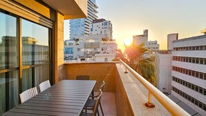 Un balcon avec une table et des chaises, d’où l’on peut admirer un paysage urbain avec ses bâtiments et un coucher de soleil.